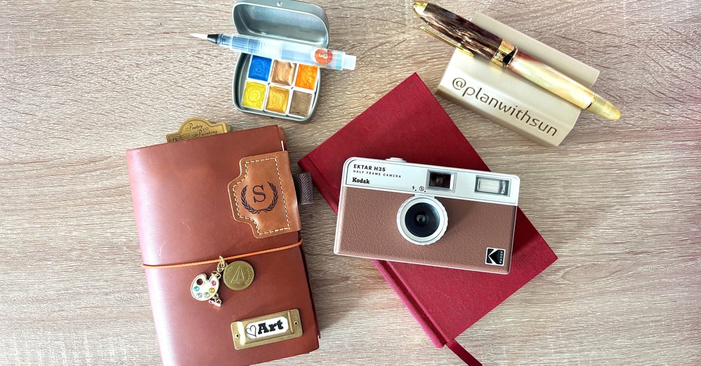 Flat lay of a leather traveler's notebook, Kodak film camera, and watercolor palette on a wooden desk.