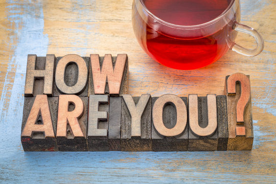 Wooden letterpress blocks spelling How Are You? next to a glass mug of red herbal tea on a rustic table.