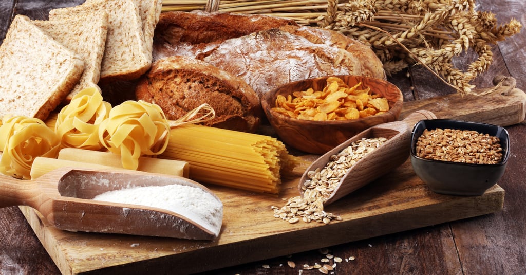 Variety of whole grain foods including bread, pasta, oats, and flour on a rustic wooden board.