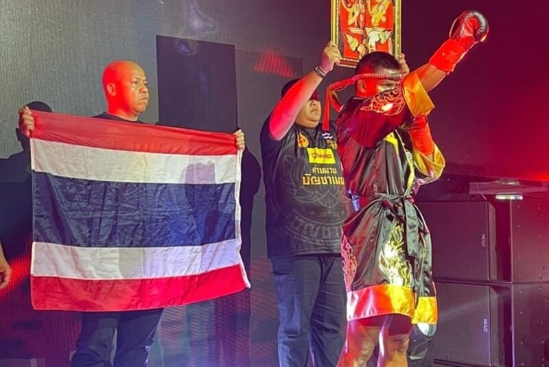 Ein Muay-Thai-Kämpfer in traditioneller Robe hält auf einer beleuchteten Bühne ein Porträt