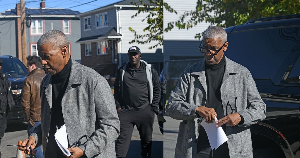 Actor Denzel Washington wearing a grey wool coat and black turtleneck holding a script on a film set.