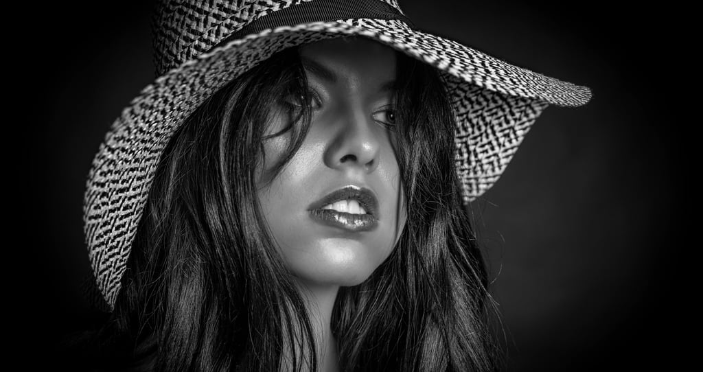 Ein Close-up-Porträt einer Frau in Schwarz-Weiß, die einen breitkrempigen Hut mit einem gemusterten Band trägt. Ihr Blick ist