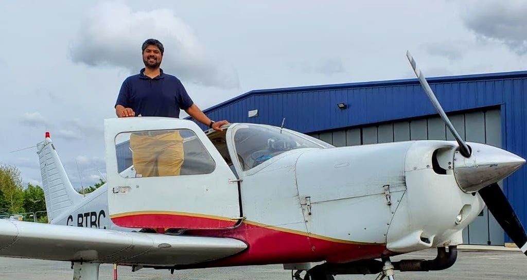 A smiling pilot standing in the cockpit of a white and red light aircraft parked near a blue hangar.