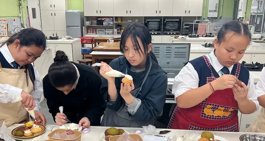 同學們喺設備齊全嘅廚房中專注裝飾甜點，使用唧花袋為 泡芙加上糖霜和士多啤梨裝飾，營造出互動學習嘅熱鬧氛圍