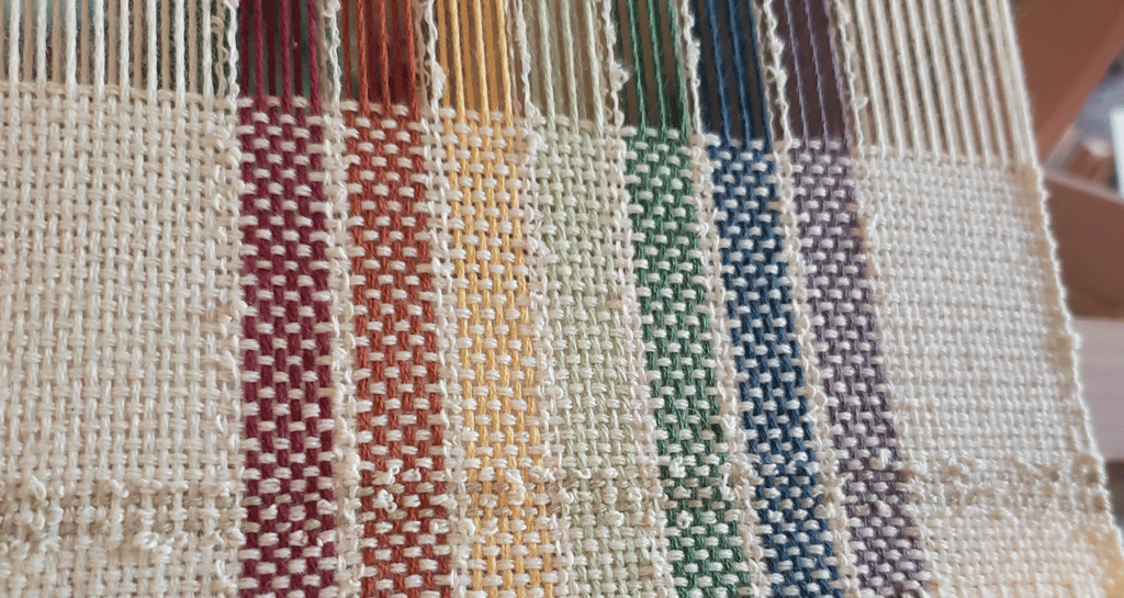 a loom with an unfinished hand-woven towel in muted rainbow pride flag colours