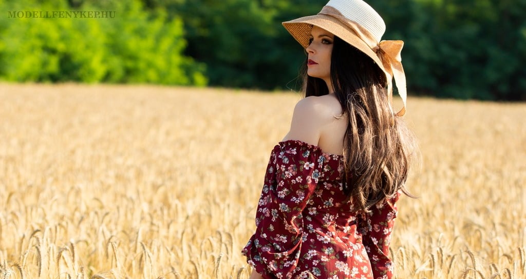 a beautiful woman in a dress and hat standing in a wheat field summertime