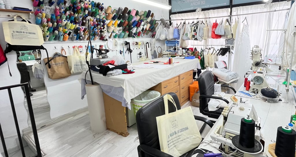 Vista general de un taller de costura con varias máquinas de coser, mesa de corte, rollos de tela, h