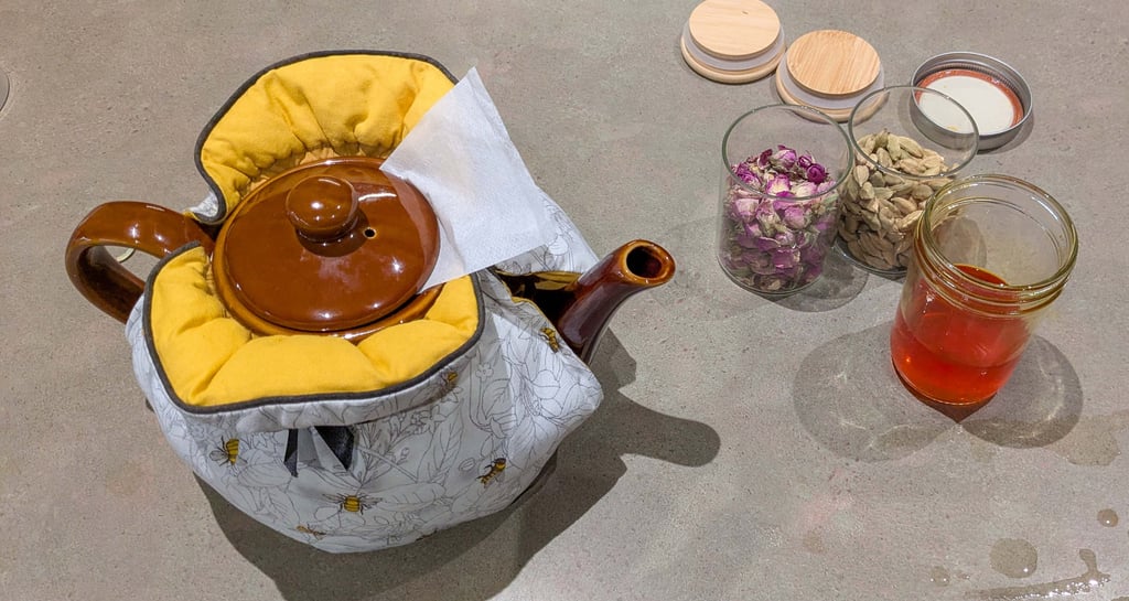 Teapot in a fabric warmer with rose buds, cardamom, and saffron on a kitchen counter.
