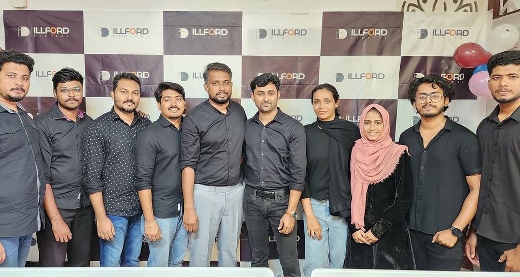 a group of men and women standing in front of a wall with a large banner - Illford Digital Academy