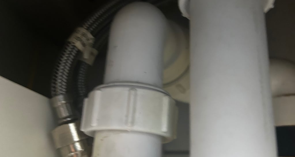 Under-sink plumbing showing a white plastic P-trap pipe and braided metal water supply line.