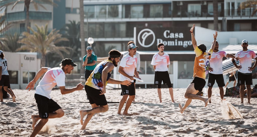 Fotografía de dos equipos compitiendo en un partido de ultimate frisbee en una playa de día