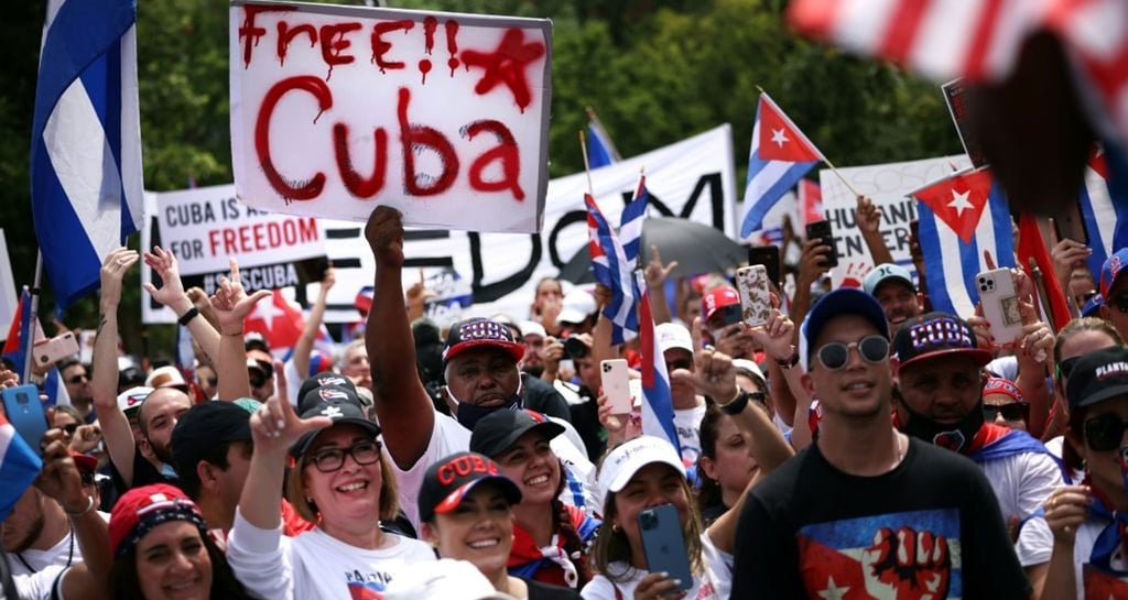 Protesters at a rally holding a Free Cuba sign and Cuban flags to support political freedom.