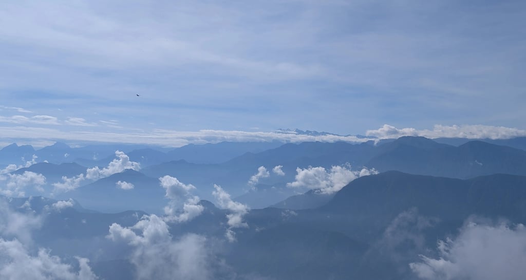Panoramic view of blue mountain ranges and misty valleys under a bright sky with wispy clouds.