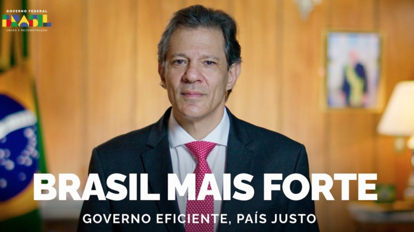 Fernando Haddad, de terno e gravata grená, em gabinete, com a Bandeira e o retrato de Lula ao fundo