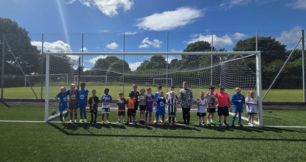A youth football team posing together in front of a goal on a sunny outdoor astro pitch.