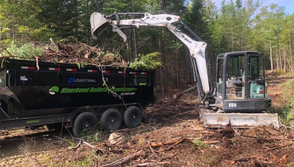 Bobcat E55 mini excavator loading tree debris into a Riverbend Rolloffs dump trailer for land clearing.