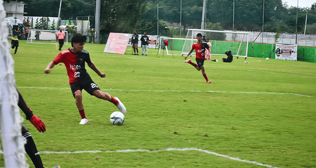 A soccer player in a red and black jersey dribbles a soccer ball on a green field during a match.