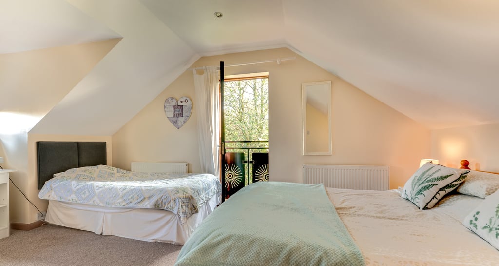 A bright attic bedroom with two comfortable beds and a balcony overlooking green trees.