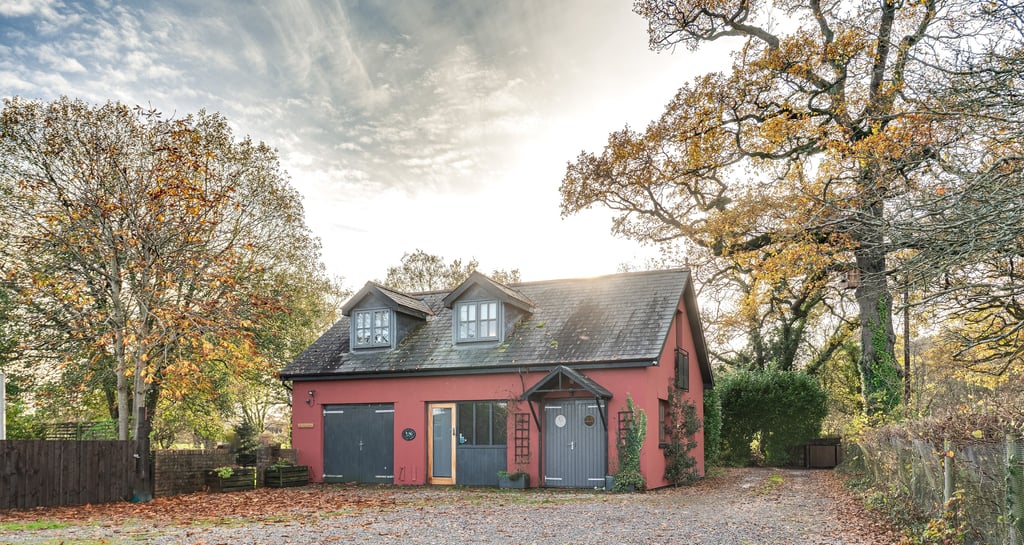 Red country cottage with grey doors and gravel driveway surrounded by autumn trees under a bright sky.