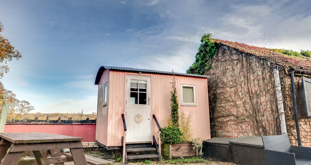 A small pink shepherd's hut with a white door next to a rustic brick outbuilding.