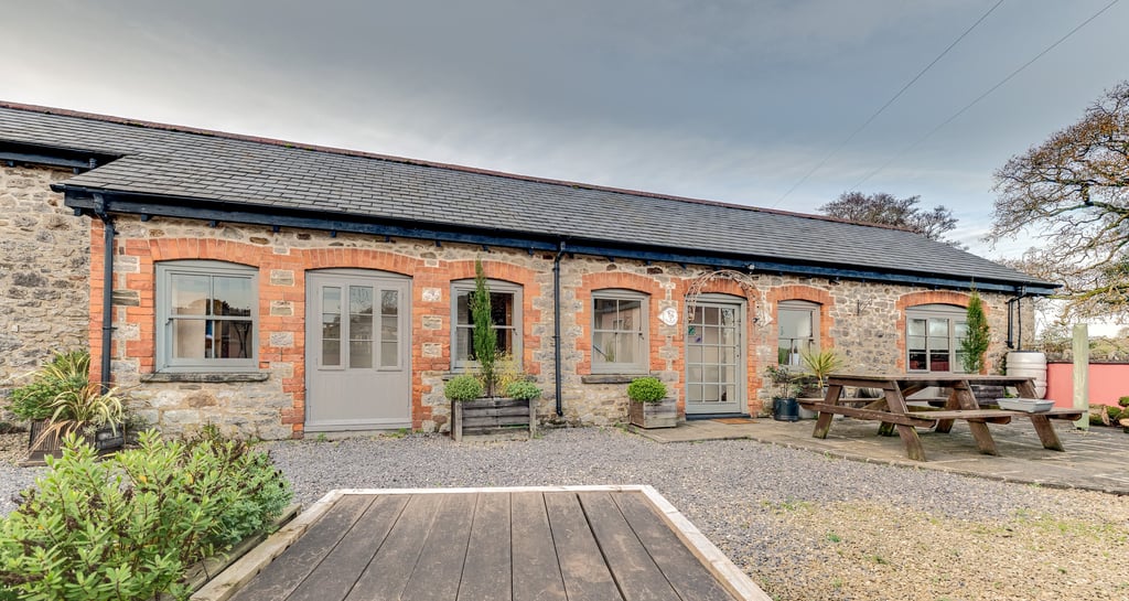 Rustic stone holiday cottage with grey doors, gravel courtyard, and wooden picnic table.