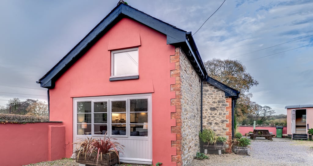 A charming pink stone cottage with a gravel driveway and outdoor seating area under a blue sky.