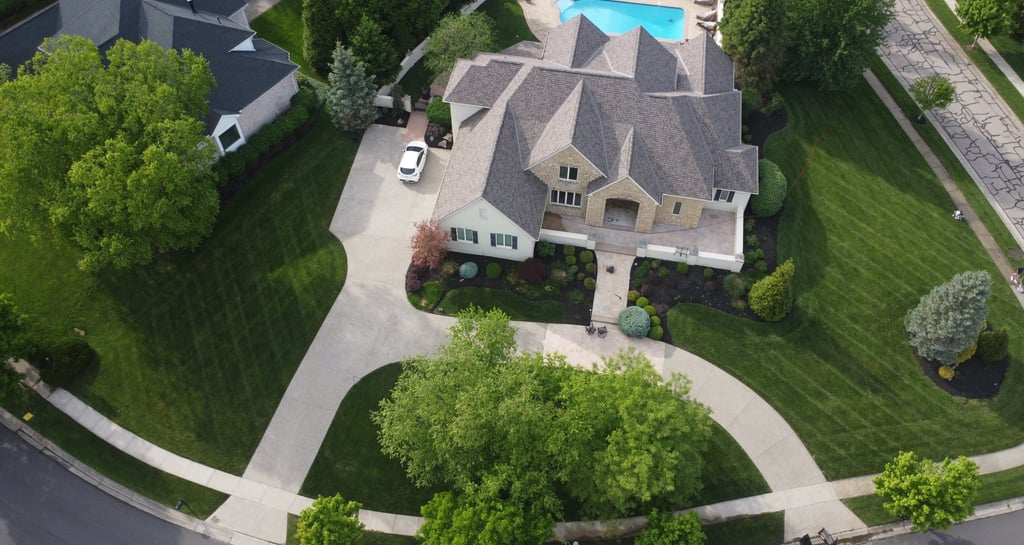 Aerial view of a luxury suburban home with a gray shingle roof, large lawn, and swimming pool.