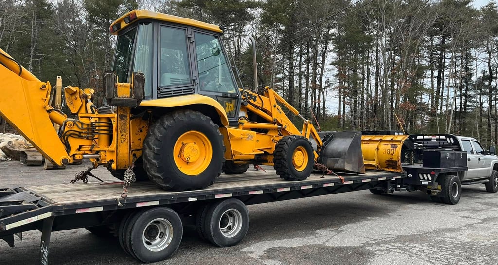 Yellow JCB backhoe loader on a flatbed trailer with a plow, ready for transport.