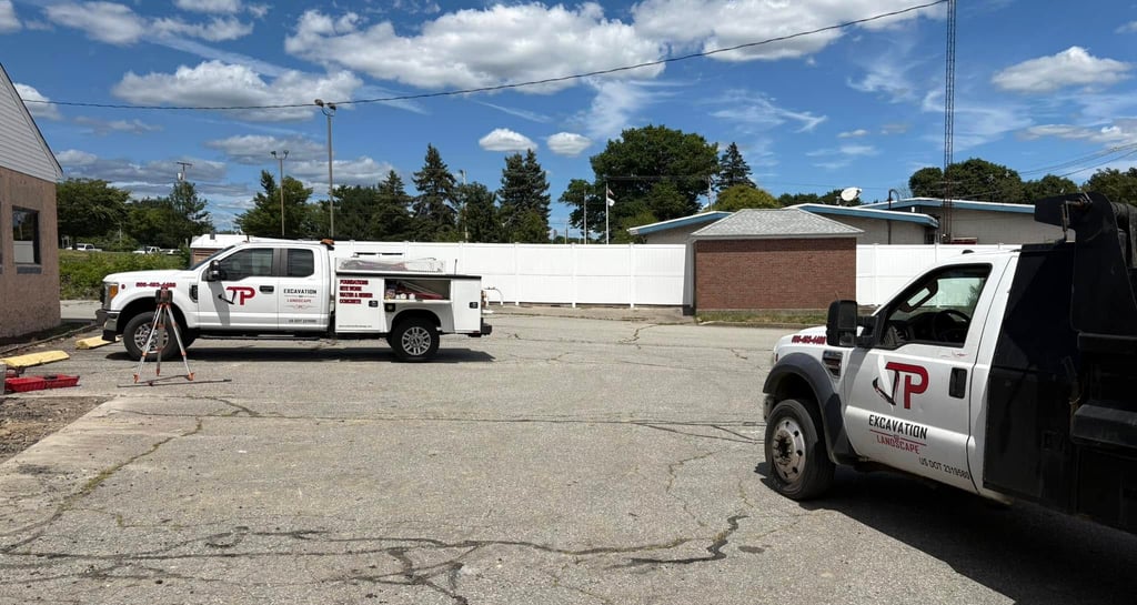 Two JP Excavation & Landscape trucks, a white service truck and a dark dump truck, parked on asphalt under a blue sky, ready