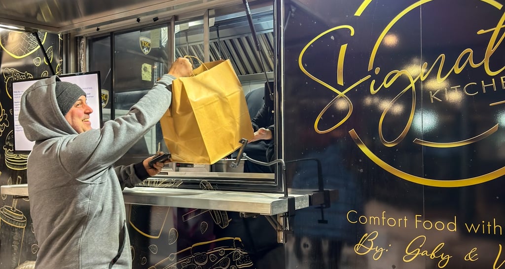 A customer picks up a takeout bag from the window of a local food truck serving comfort food at night.
