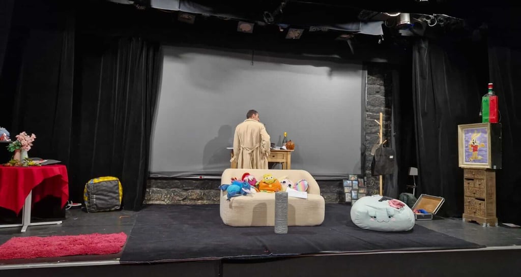 Scène de théâtre en décor domestique : une personne en robe beige est debout à une table, entourée d