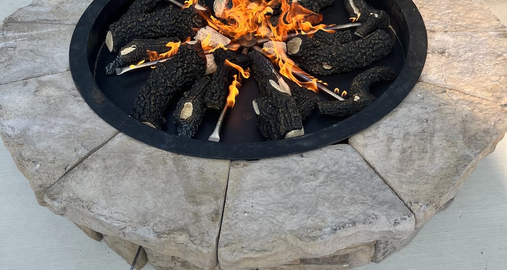 A circular outdoor stone gas fire pit with burning logs on a concrete patio.