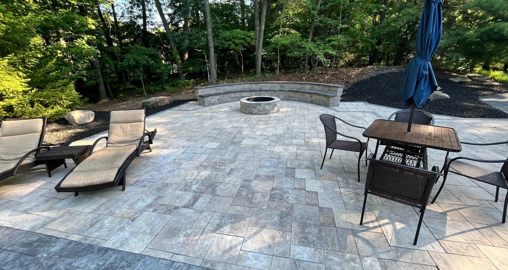 A backyard stone paver patio featuring a fire pit, curved stone bench, and outdoor dining set with lounge chairs.