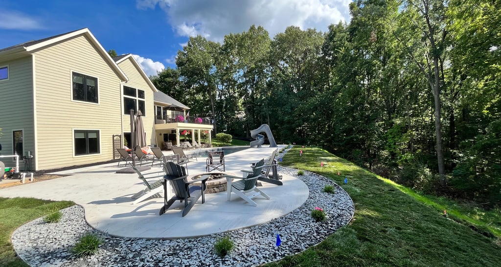 Luxury backyard concrete patio with fire pit, lounge chairs, and pool slide bordering a lush green forest.
