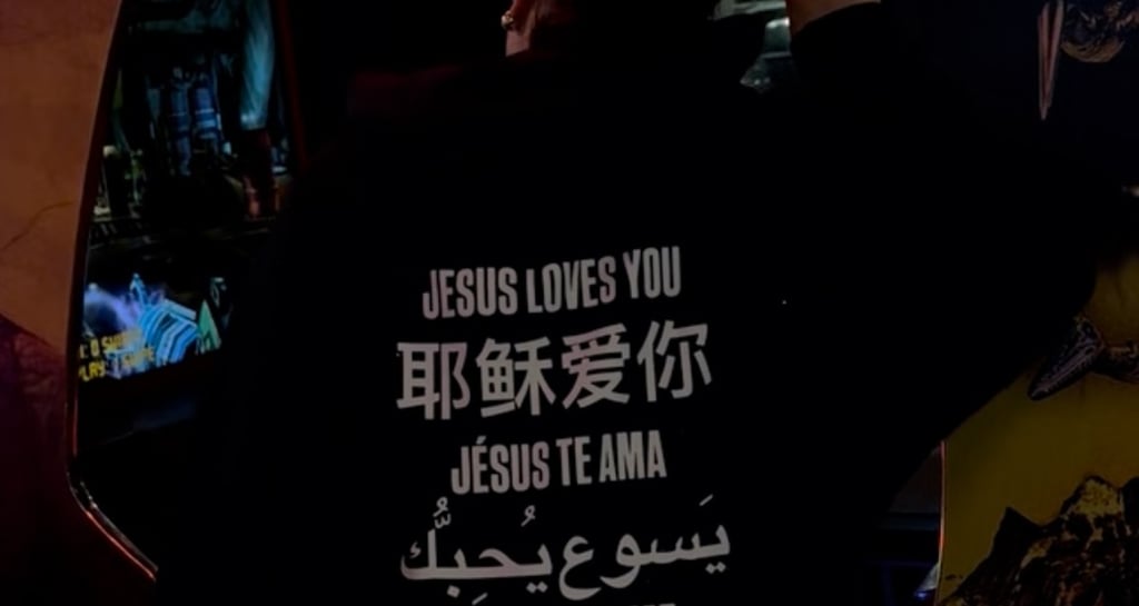 Back view of person in hoodie with 'Jesus loves you' in many languages, arms raised in neon arcade.