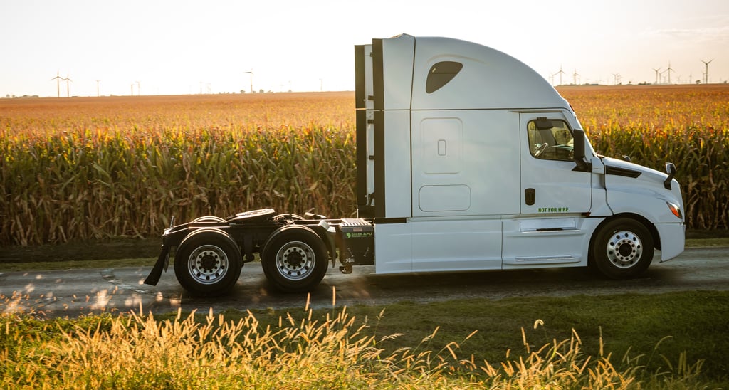 A white semi-truck tractor driving past a sunlit cornfield with wind turbines in the background.