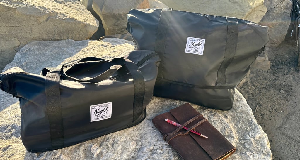 Two black Night Supply Co. duffel bags on rocks beside leather journal and pen.