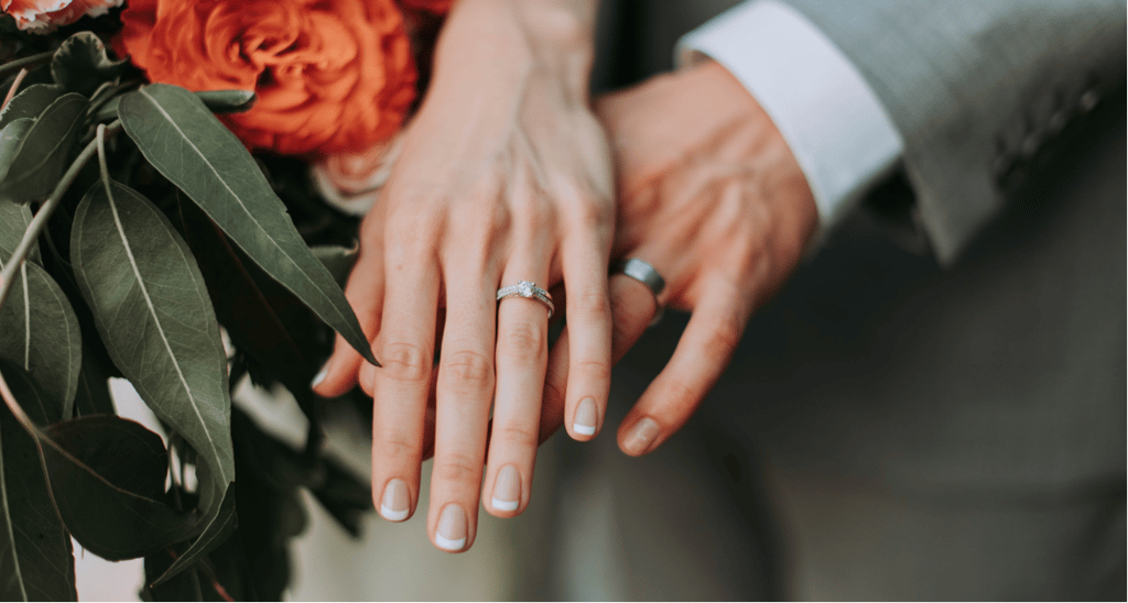 Close-up of a married couple's hands displaying a diamond engagement ring and wedding bands next to a bridal bouquet.