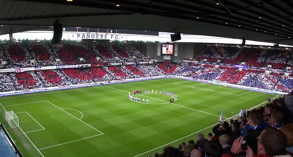 Innenansicht des Ibrox Stadium mit grünem Spielfeld und blauen Sitzreihen.