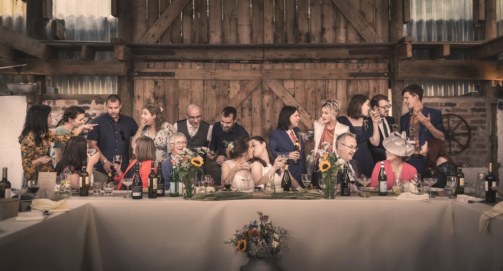 Wedding party celebration at a rustic barn reception with guests toasting at a long table.
