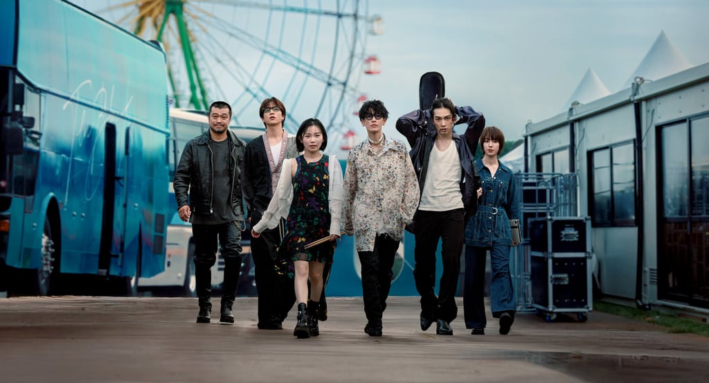 A stylish Japanese rock band walking outdoors past a tour bus and Ferris wheel at a festival venue.