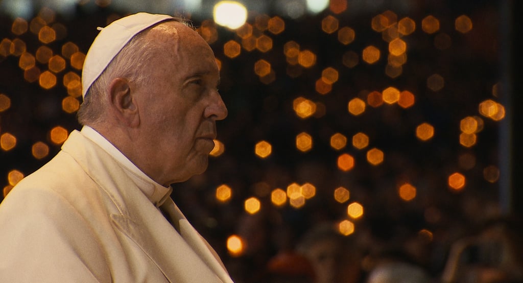 Pope Francis in profile at night with golden bokeh candle lights in the background.