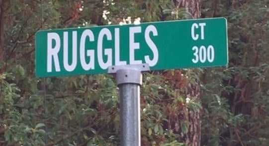 Photo of green street sign in front of wooded background: "Ruggles Ct, 300."