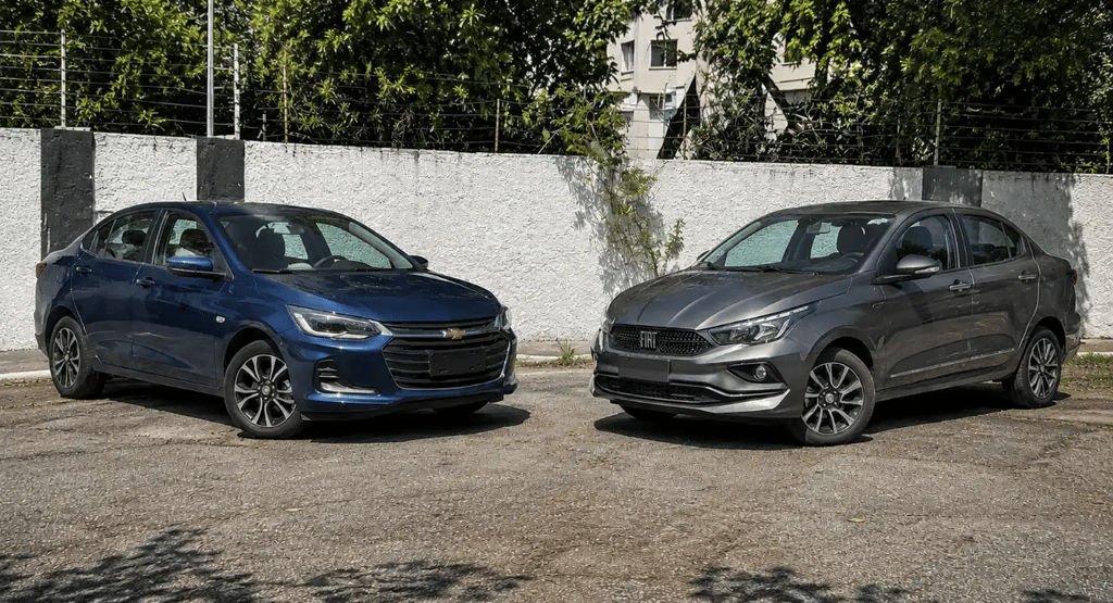 Chevrolet Onix Plus e Fiat Cronos em perspectiva 3/4, posicionados frente a frente.