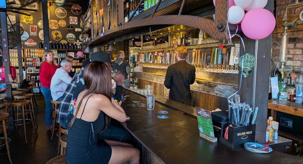 Customers sit at a rustic wooden bar in a craft brewery with many draft beer taps and industrial decor.