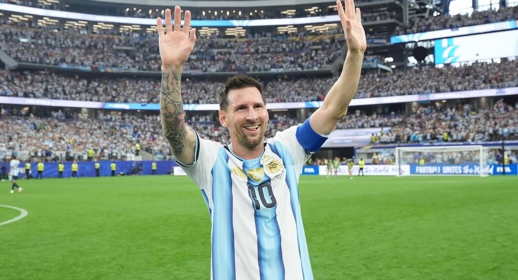 Lionel Messi waving to fans in an Argentina jersey at a crowded soccer stadium.