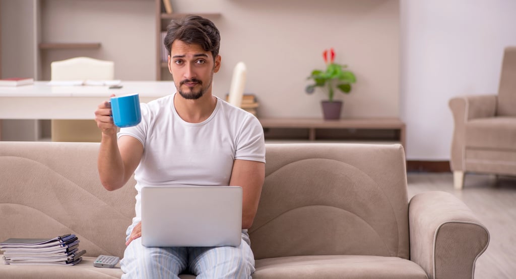 A man in pajamas sitting on a couch working on a laptop while holding a blue coffee mug.