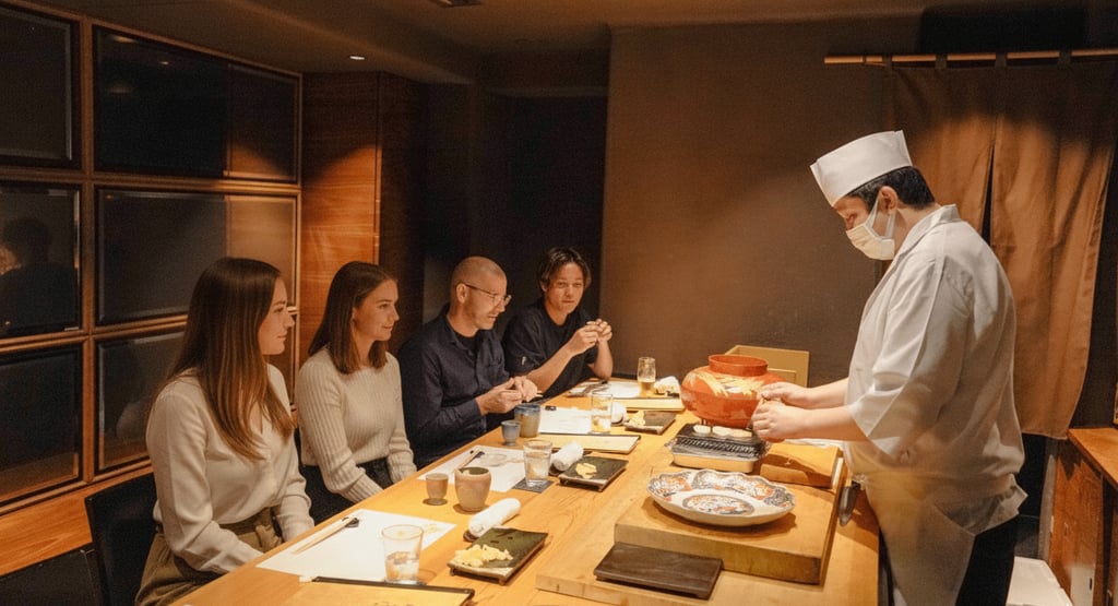 A chef in a white uniform and face mask serves food to four seated people at a wooden table with dis