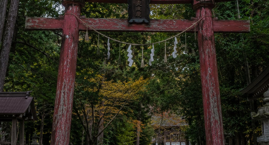 A weathered red wooden torii gate marks the entrance to a traditional Japanese Shinto shrine in a lush forest.