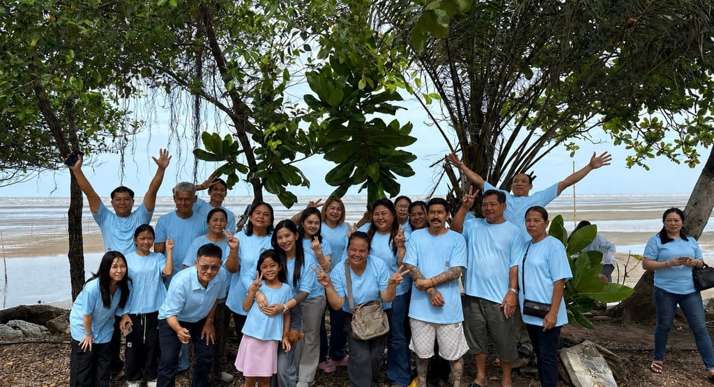 A large group of people in matching light blue t-shirts posing for a photo outdoors near a beach with trees.